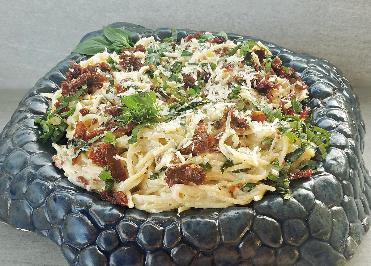 Side view of pasta tossed with sun-dried tomatoes, parmesan, garnished with fresh basil, served on a highly textured bowl.