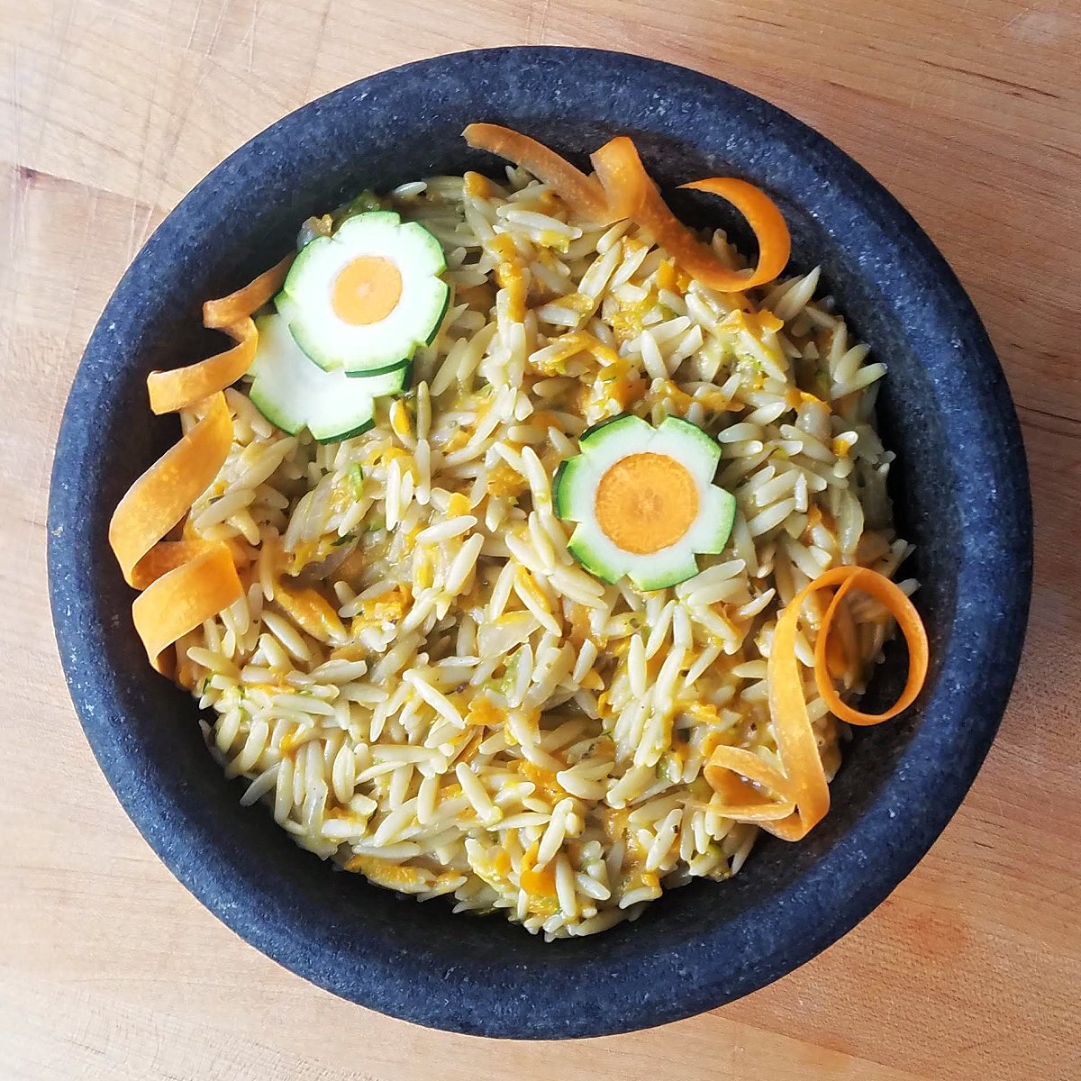 Overhead shot of cooked orzo in a stone bowl, garnished with zucchini-carrot flowers and carrot curls. 