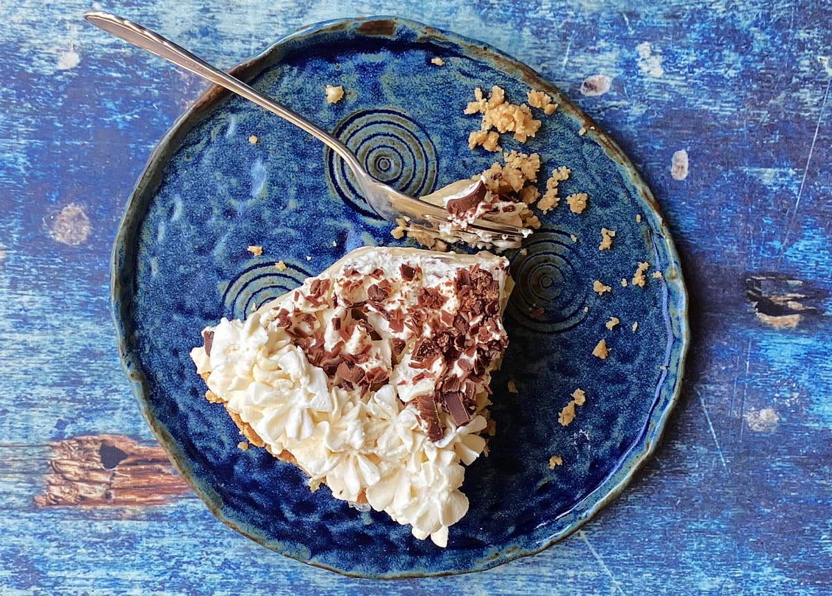 Overhead shot of a slice of whipped cream topped butterscotch pie on a hand-built plate. A fork has taken off one bite. 