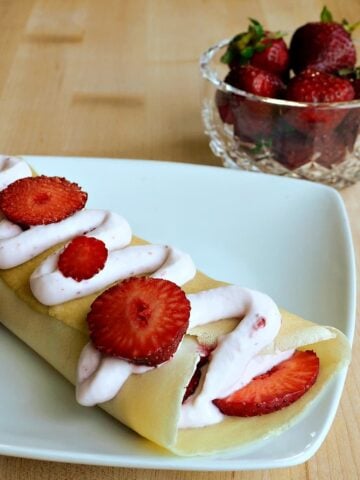 One crepe filled with fresh strawberries, with a zigzag of strawberry cream and more fresh strawberries on top. Bowl of strawberries in the background.