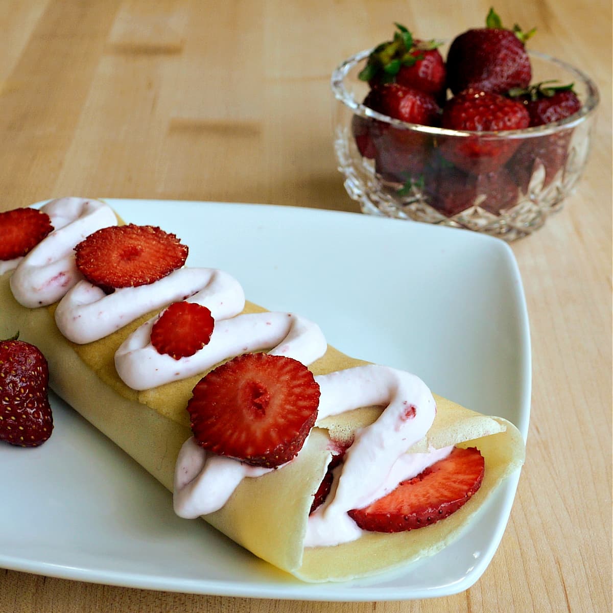 One crepe filled with fresh strawberries, with a zigzag of strawberry cream and more fresh strawberries on top. Bowl of strawberries in the background.