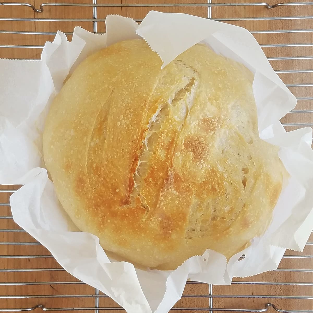 Boule of baked sourdough bread setting on a large square of parchment that has been gathered a bit around the bottom of the loaf.