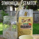 Jar of prepared sourdough starter, with wooden spoon sticking out. Small yellow teacup marked with ½ cup, and a bag of rye flour.