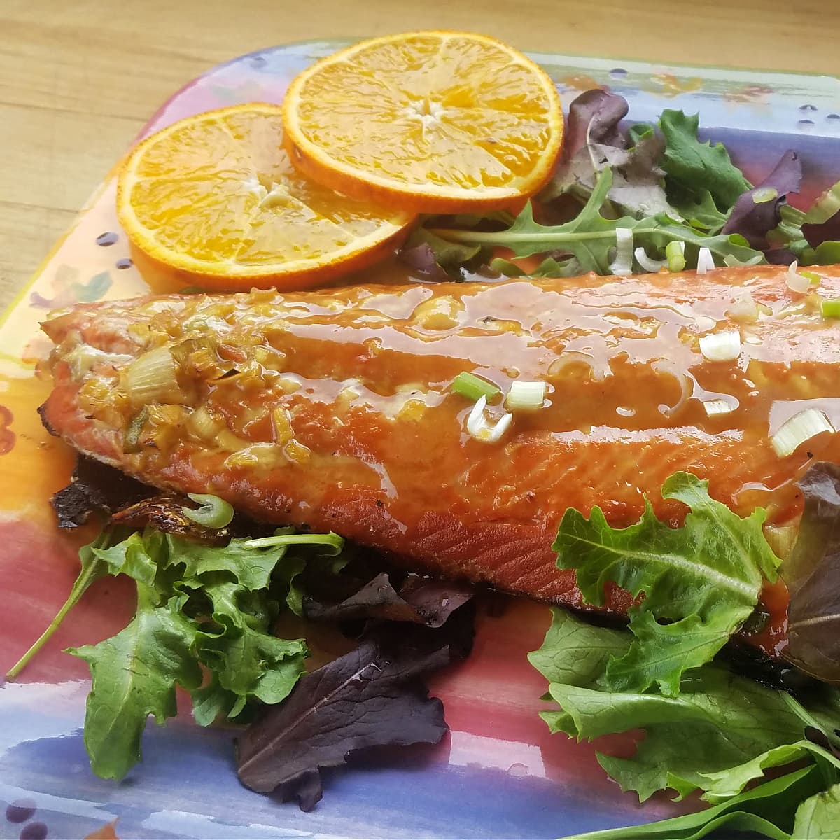 Large grilled salmon fillet on a bed of greens on a colorful rectangular platter, garnished with orange slices.