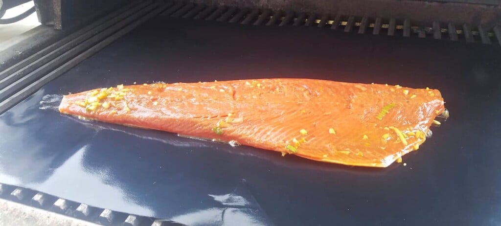 Uncooked salmon fillet on black grill mat, on top of BBQ grill. 