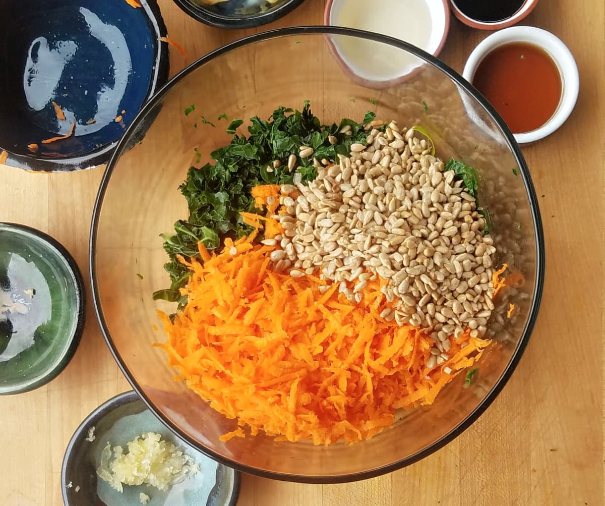 Shredded carrots, sunflower seeds, kale, and other ingredients in large glass bowl, unmixed.