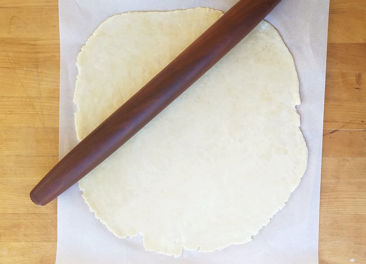 A french rolling pin angled across a rolled out piece of pie dough, on parchment.