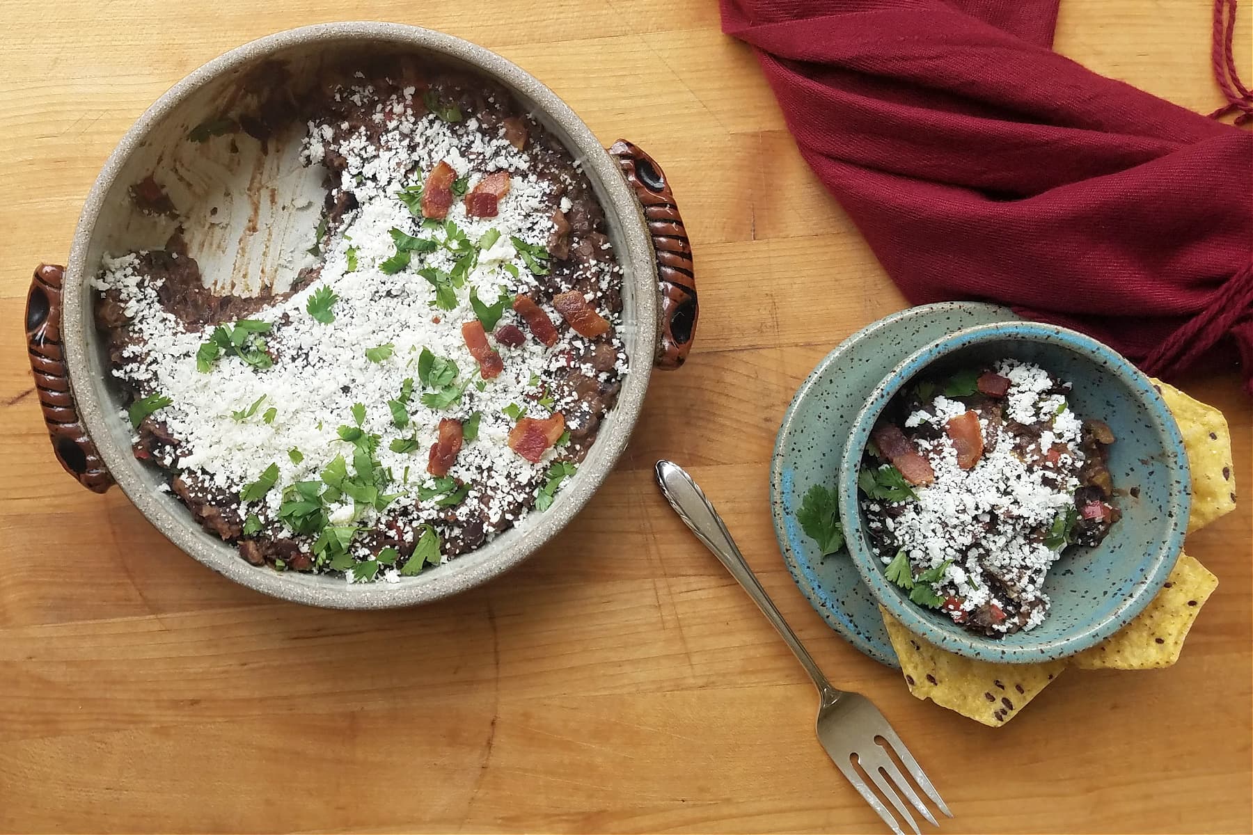 Round crockery casserole dish filled with cotija covered black beans, with one portion removed. Small pottery bowl with portion of black beans. Fork to the side.