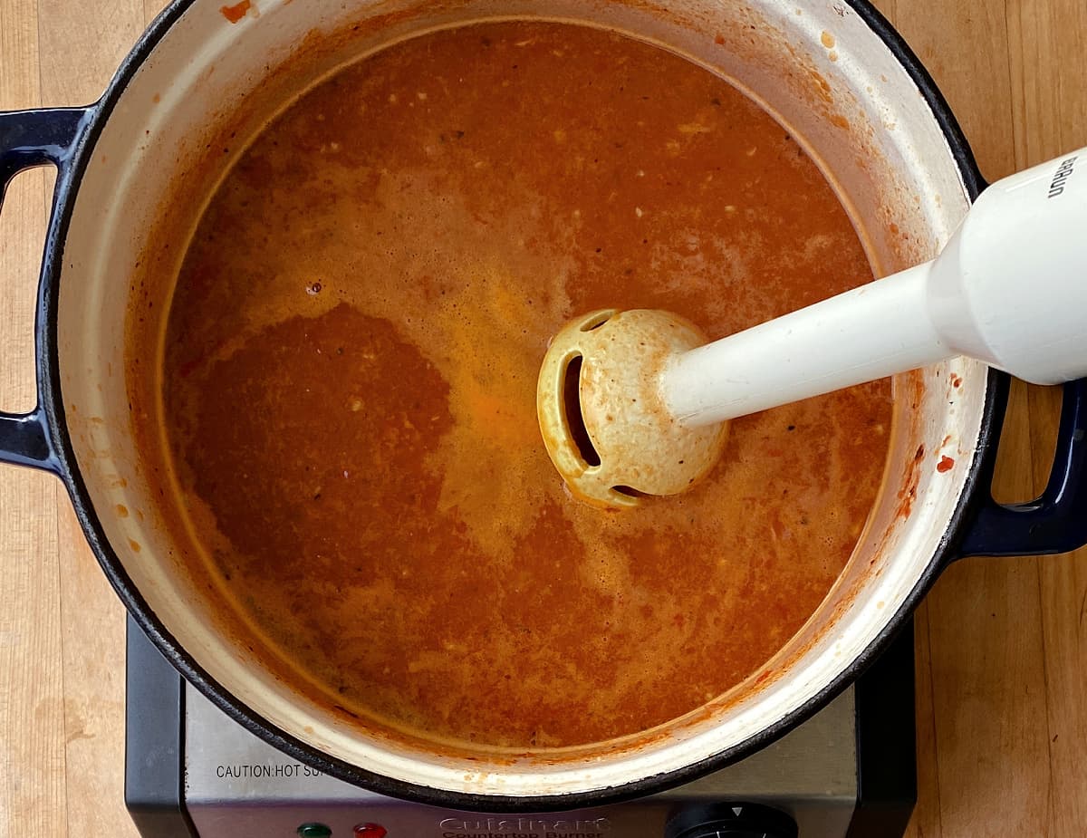 Immersion blender submerged in red enchilada mixture in Dutch oven. 
