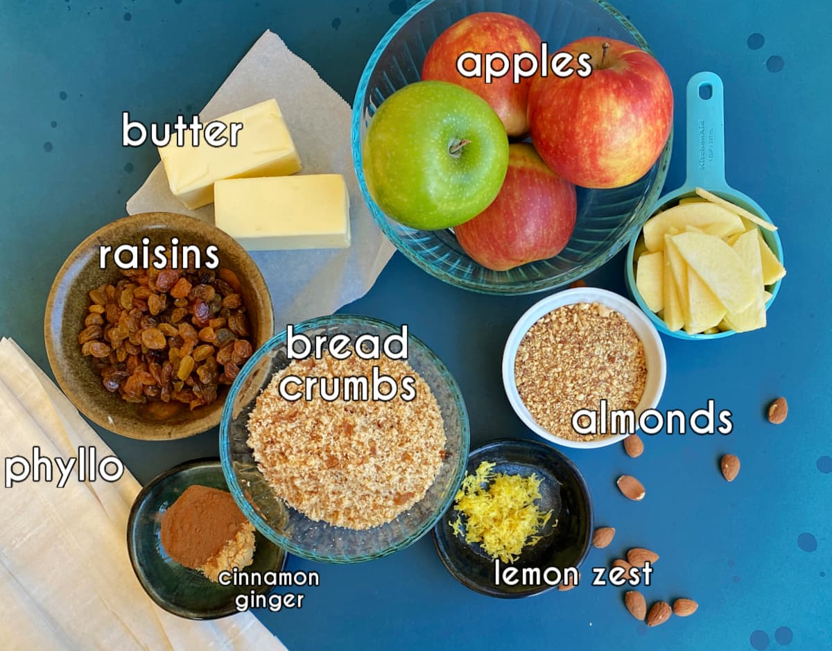 Apple strudel ingredients, labeled: butter, apples, almonds, lemon zest, bread crumbs, raisins, phyllo, spices.