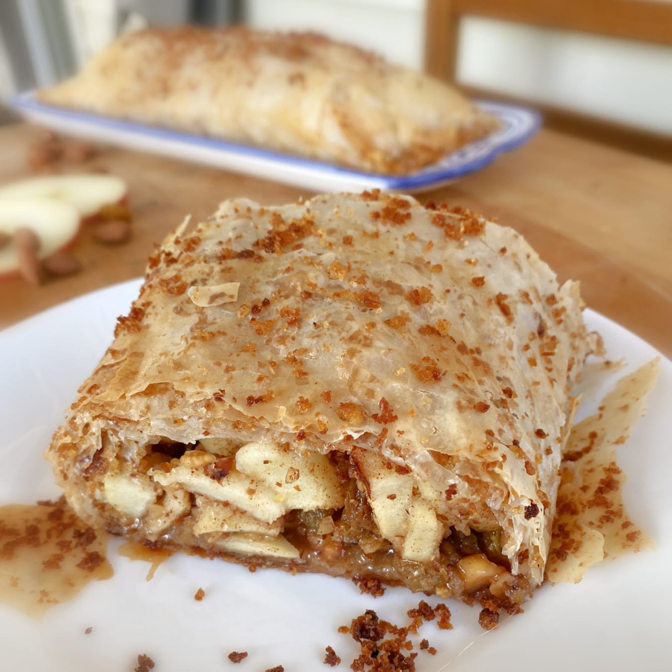 Cut piece of apple strudel on small plate, with a roll of strudel in the background.