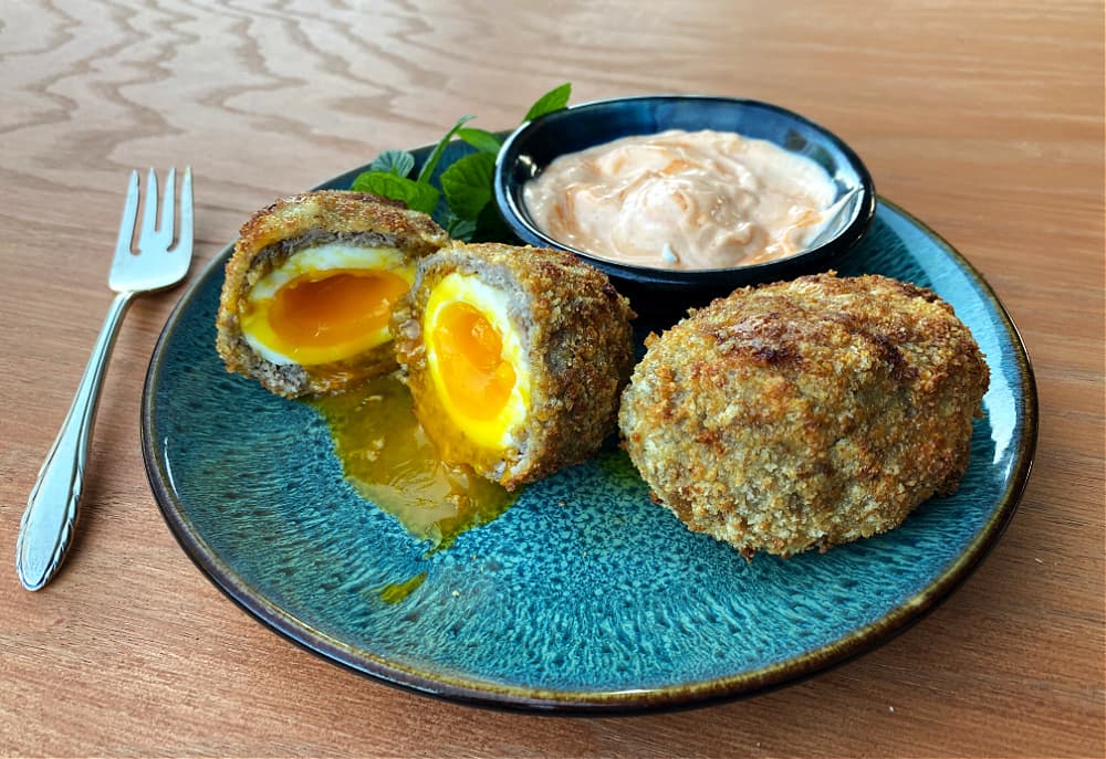 Two Scotch Eggs plated with sauce, one cut open and one still whole. 