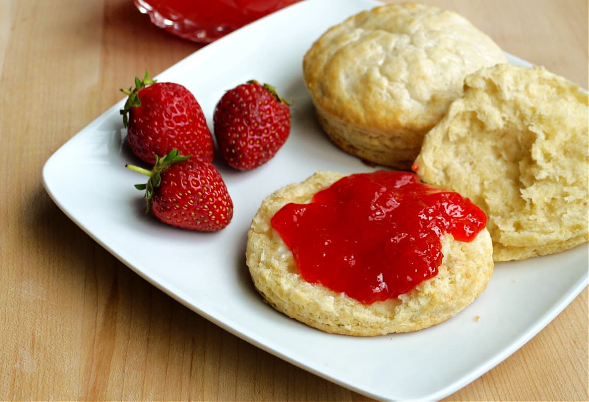 Two biscuits on a plate, with three strawberries on the side. One biscuit is sliced open and spread with strawberry jam.