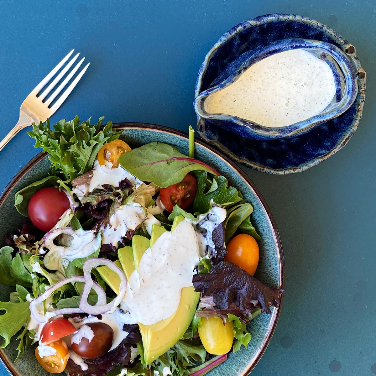 Salad dressing in a small, hand-built clay pitcher beside a bowl of salad greens, tomato, and avocado that is topped with more dressing. 