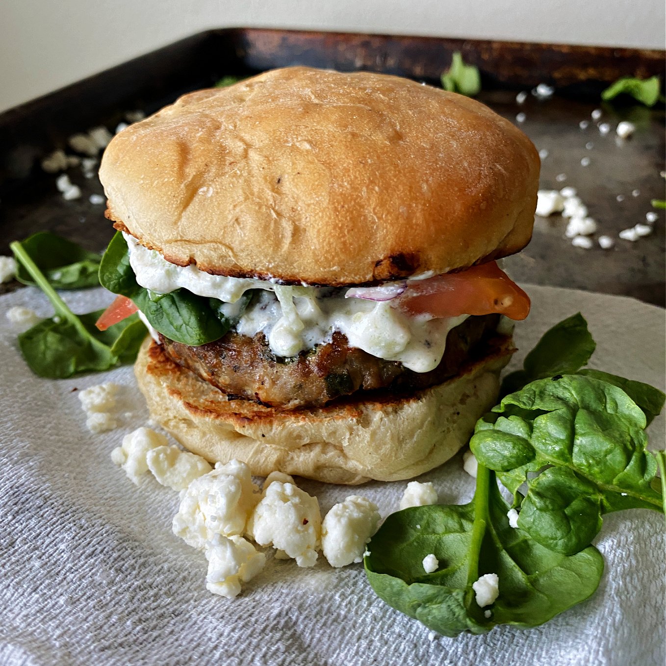 Close-up of a Greek Turkey Burger on a tray, with spinach leaves, feta around base. 