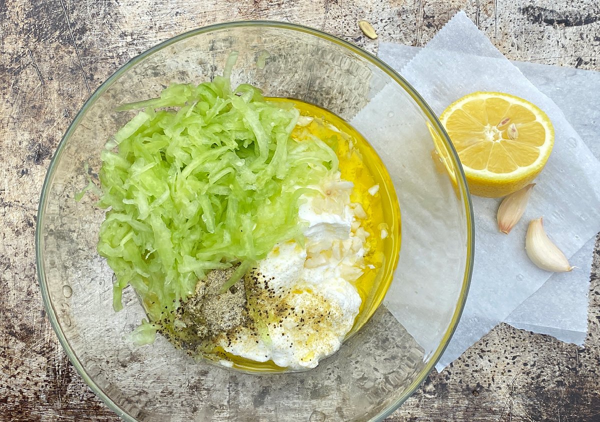 Tzadiki ingredients unmixed in glass bowl. Half a lemon and two garlic cloves on parchment square. 