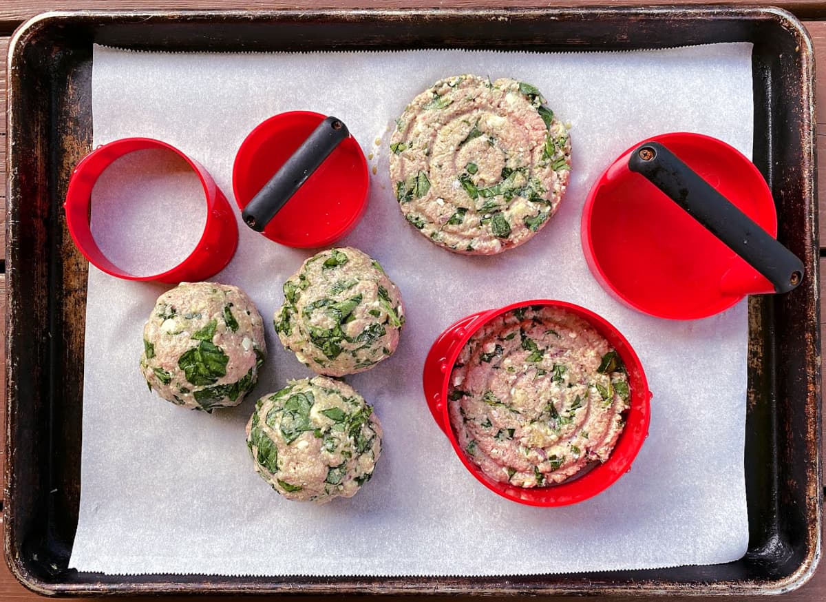 Red burger press on parchment covered old baking tray with turkey burgers and balls of burger mixture.  