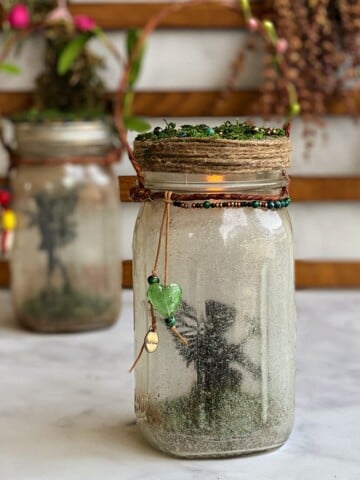Mason jar with a silhouette of a fairy inside, lighted from the inside, a jute-wrapped wire bale on top, and jute wrapped around the edge of the lid. Another fairy lantern in in the background.