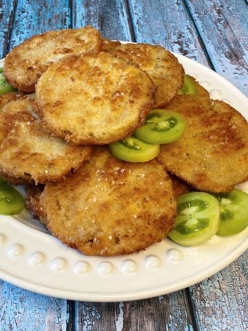 Fried green tomatoes stacked ona white plate, garnished with small slices of raw green tomatoes.