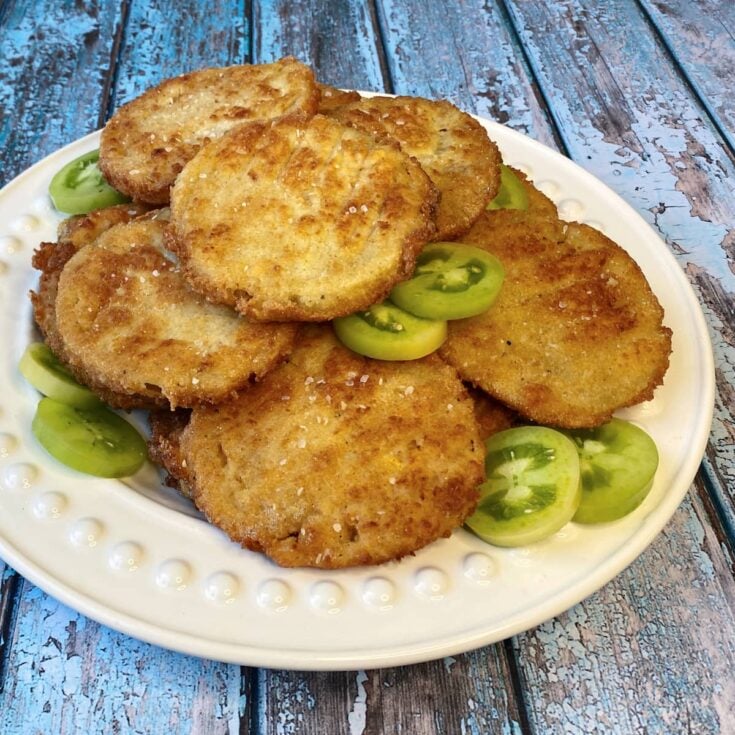Fried green tomatoes stacked ona white plate, garnished with small slices of raw green tomatoes.