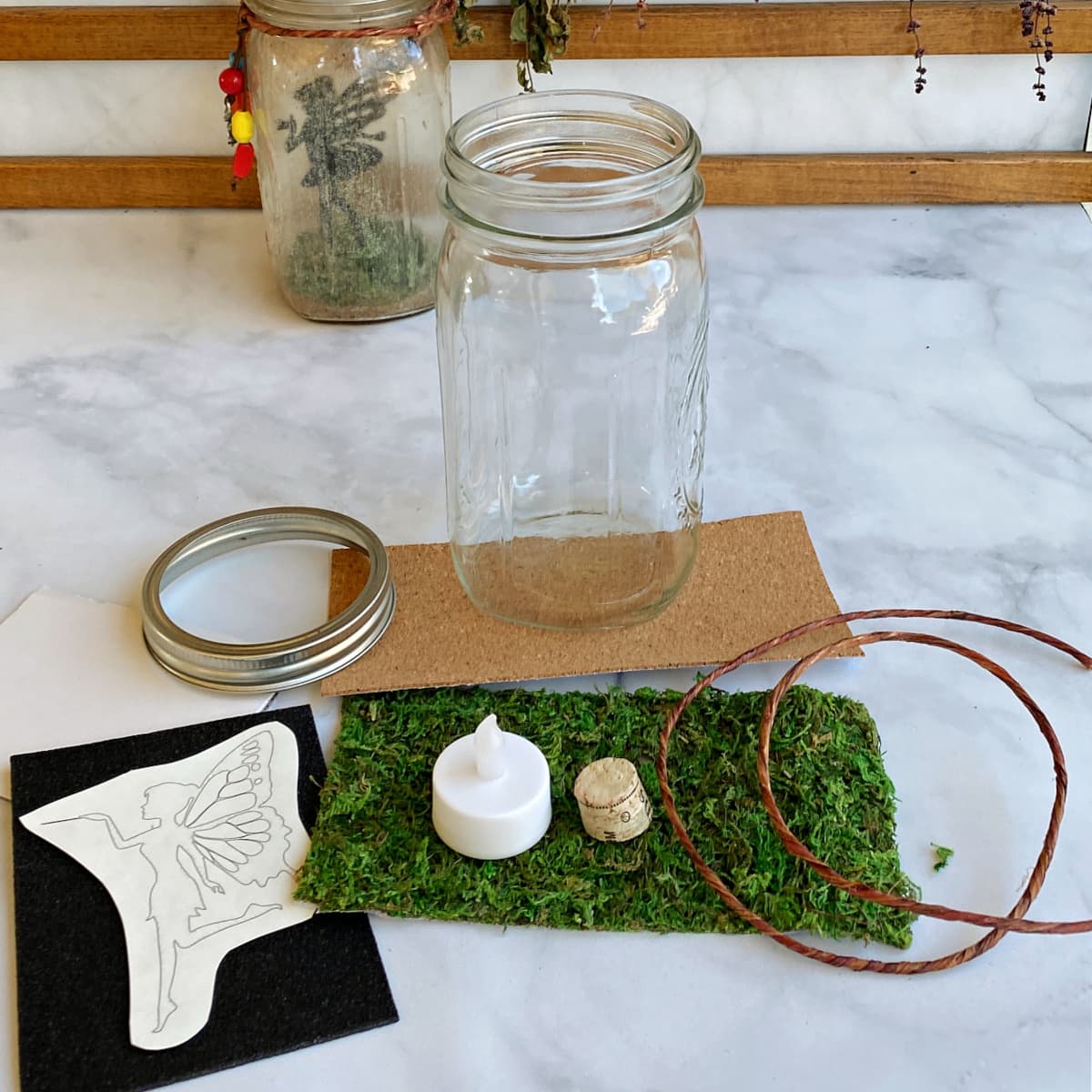 Basic materials needed for fairy jars: Mason jar and ring, fairy printout on adhesive paper, wire for bale, mini battery-powered candle, small piece of cork, black foan sheet, craft moss sheet, adhesive cork sheet.