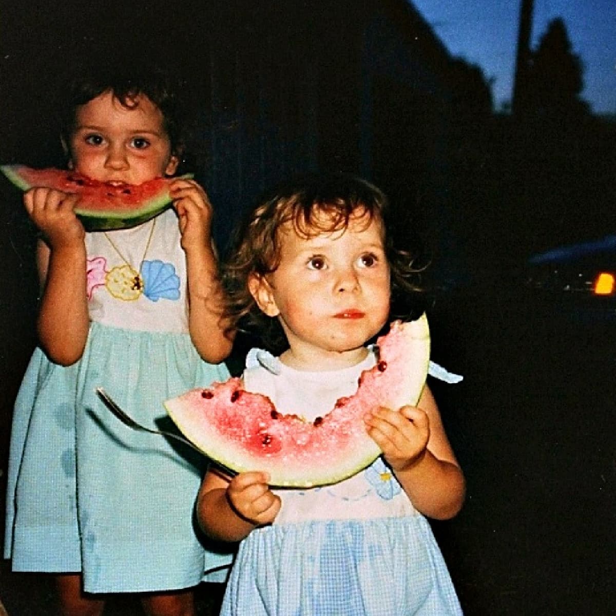 Two preschool-age girls eating watermelon slices outside at night. 