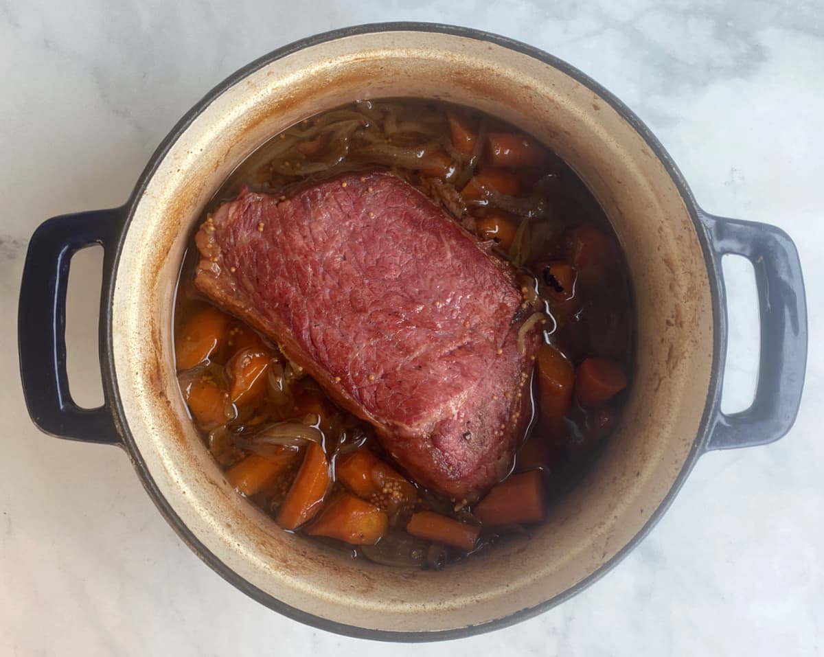 Braised corned beef in enameled Dutch oven, surrounded by cooked carrots and onions.