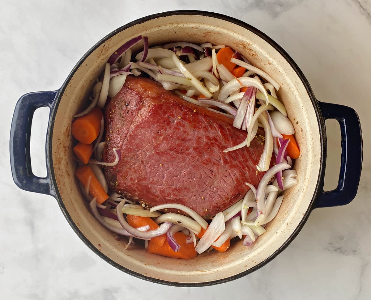 Seared corned beef in Dutch oven, surrounded by sliced onions and carrots.