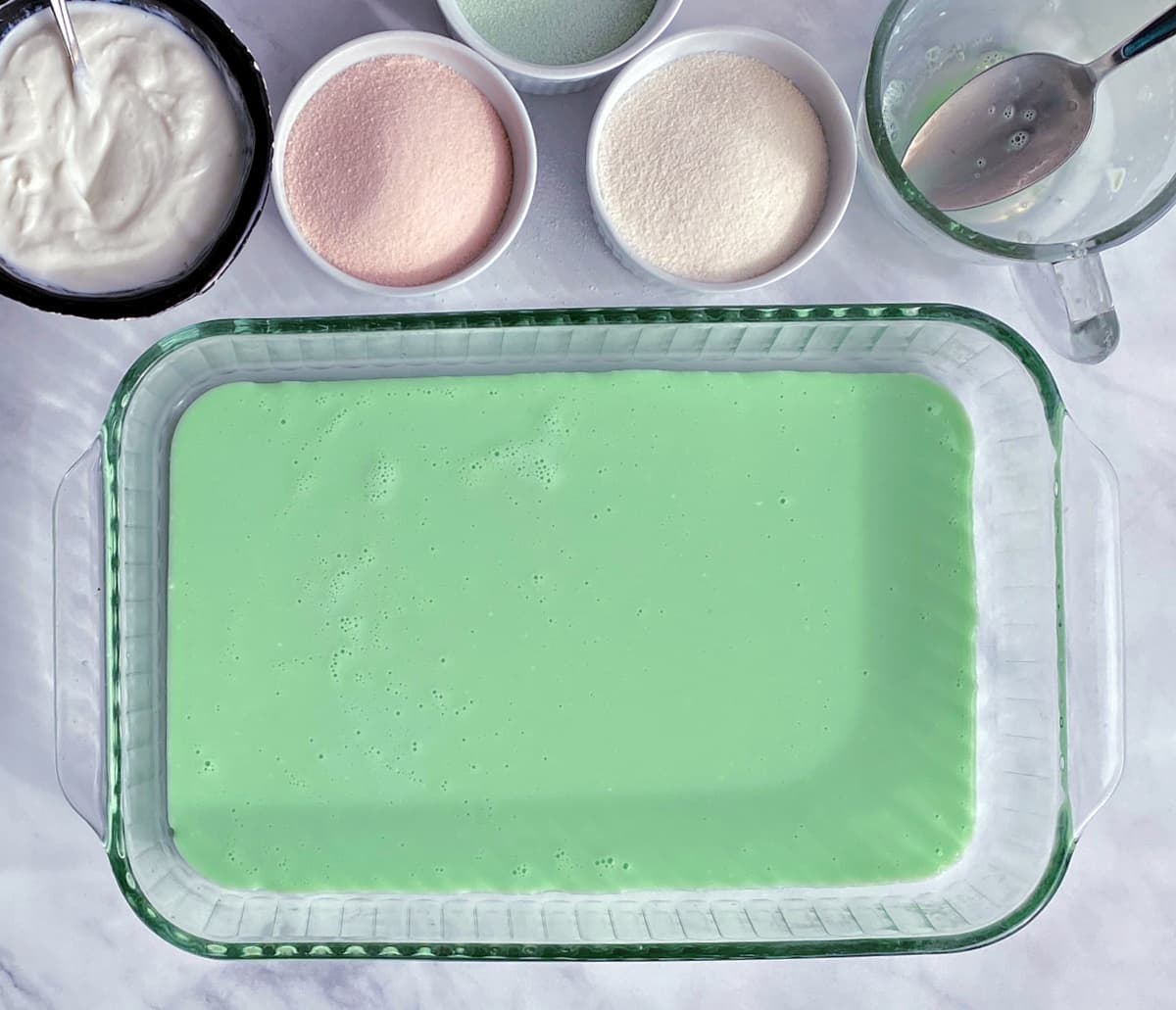 pastel green jello layer in pyrex casserole dish. Dry gelatin mix and yogurt in bowls at top of shot.
