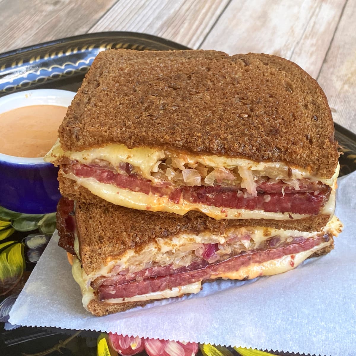 Two halves of a Rueben stacked on parchment, on a Russian black-lacquered tray. Russian dressing in a small service cup in the background.