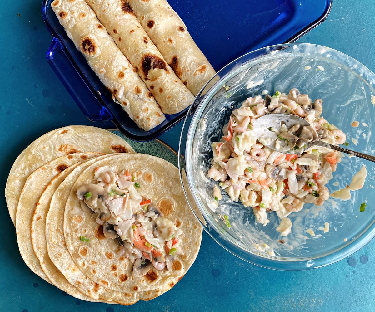 Glass bowl filled with seafood mixture, three rolled tortillas in casserole dish, and seafood mix on a tortilla, ready to roll.