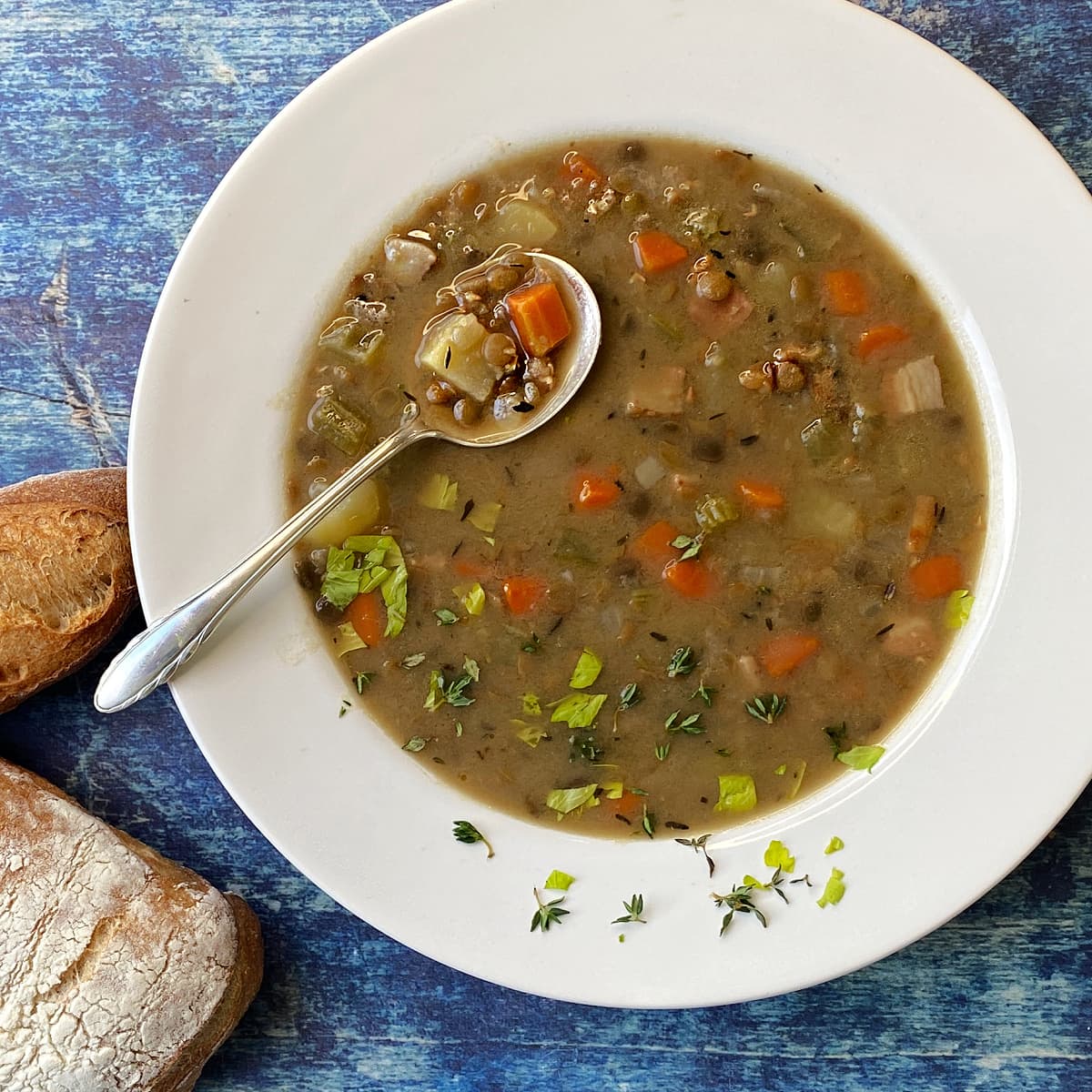 Overhead shot of a wide brimmed bowl filled with lentil soup, garnished with fresh thyme and celery leaves. Spoonful of soup. Crusty bread on the side.  
