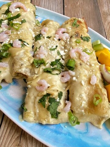 Four creamy seafood enchiladas on a platter, garnished with bay shrimp, cilantro, and cotija. Mango salad on the side.