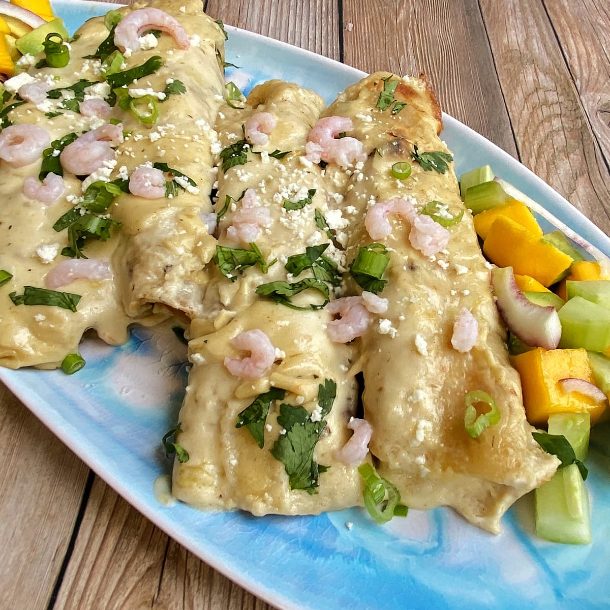 Four creamy seafood enchiladas on a platter, garnished with bay shrimp, cilantro, and cotija. Mango salad on the side.  