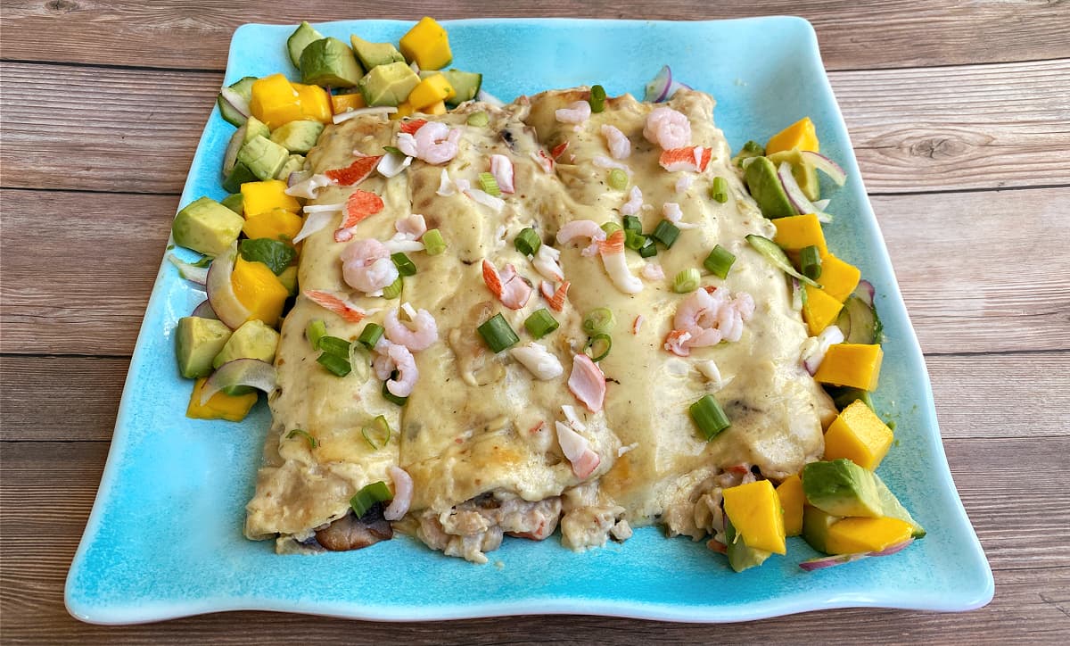 Four seafood enchiladas on a long platter, garnished with bay shrimp, green onions, and imitation crab. Mango salad on the side.
