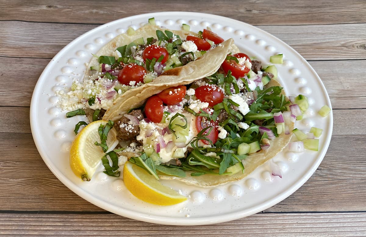 Side view of two Greek tacos: two medium flour tortillas on a plate, filled with seasoned loose meat, arugula, feta, tomatoes, yogurt, basil ribbons; garnished with lemon slices.