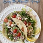 Two medium flour tortillas on a plate, filled with seasoned loose meat, arugula, feta, tomatoes, yogurt, basil ribbons; garnished with lemon slices. Pin text overlay reads: Greek Tacos | Easy 30-Minute Prep!