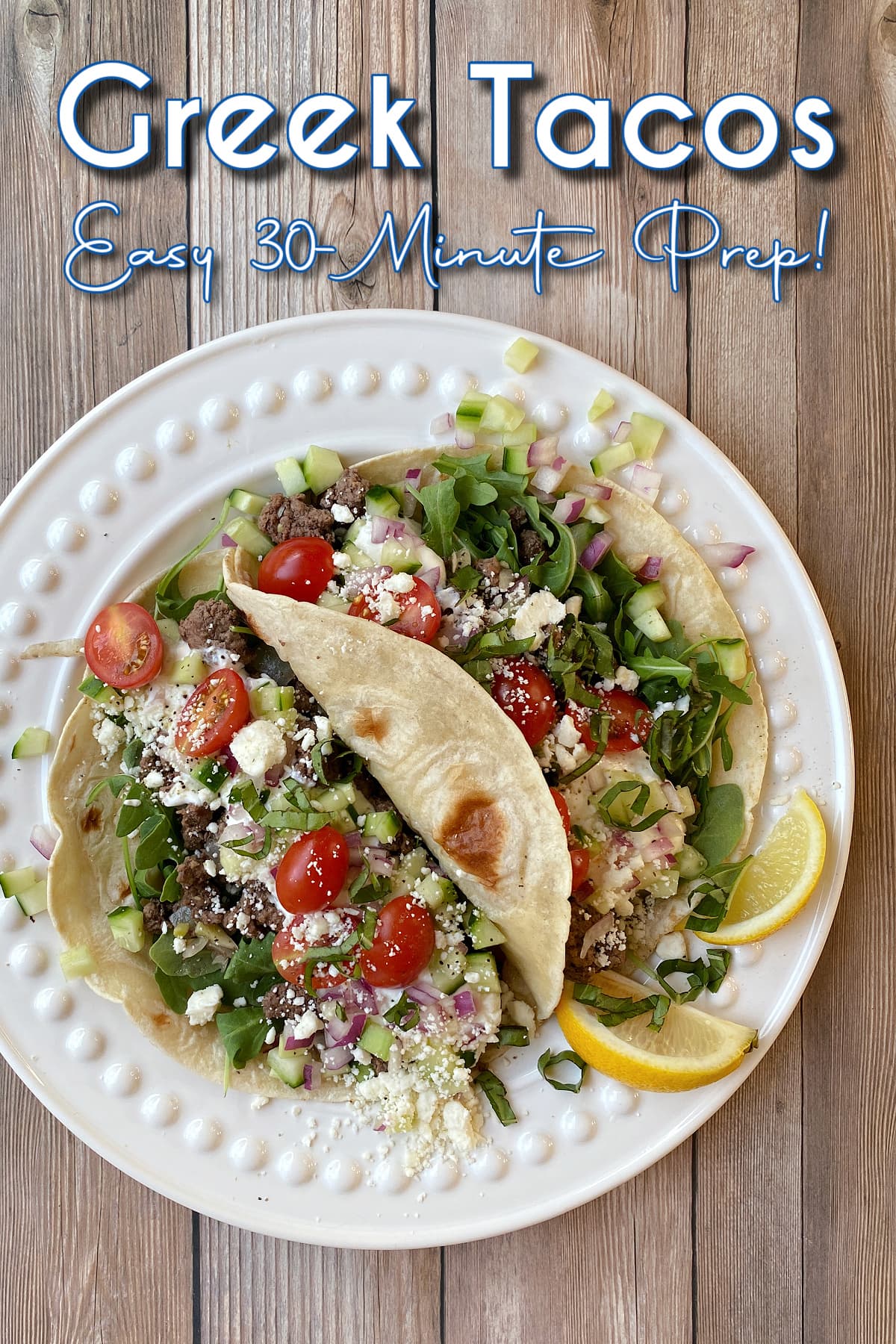 Two medium flour tortillas on a plate, filled with seasoned loose meat, arugula, feta, tomatoes, yogurt, basil ribbons; garnished with lemon slices. Pin text overlay reads: Greek Tacos | Easy 30-Minute Prep!