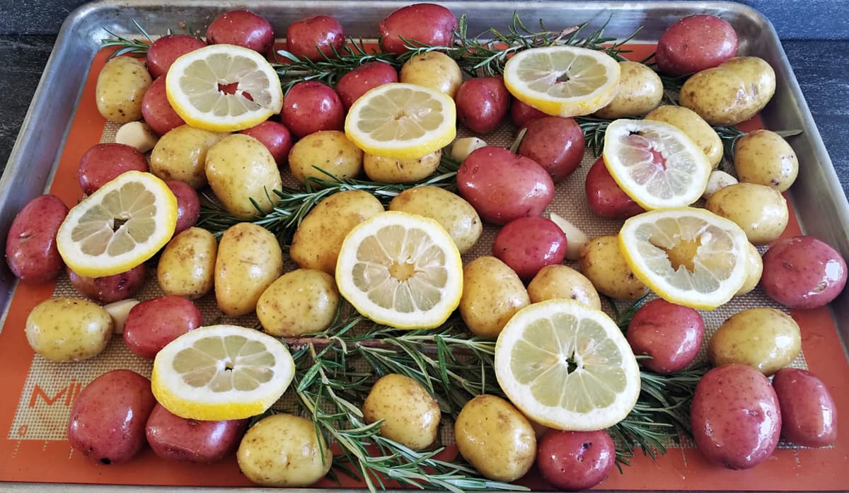 Side view of baking tray covered with baby red and Yukon gold potatoes, fresh rosemary, and lemon slices.
