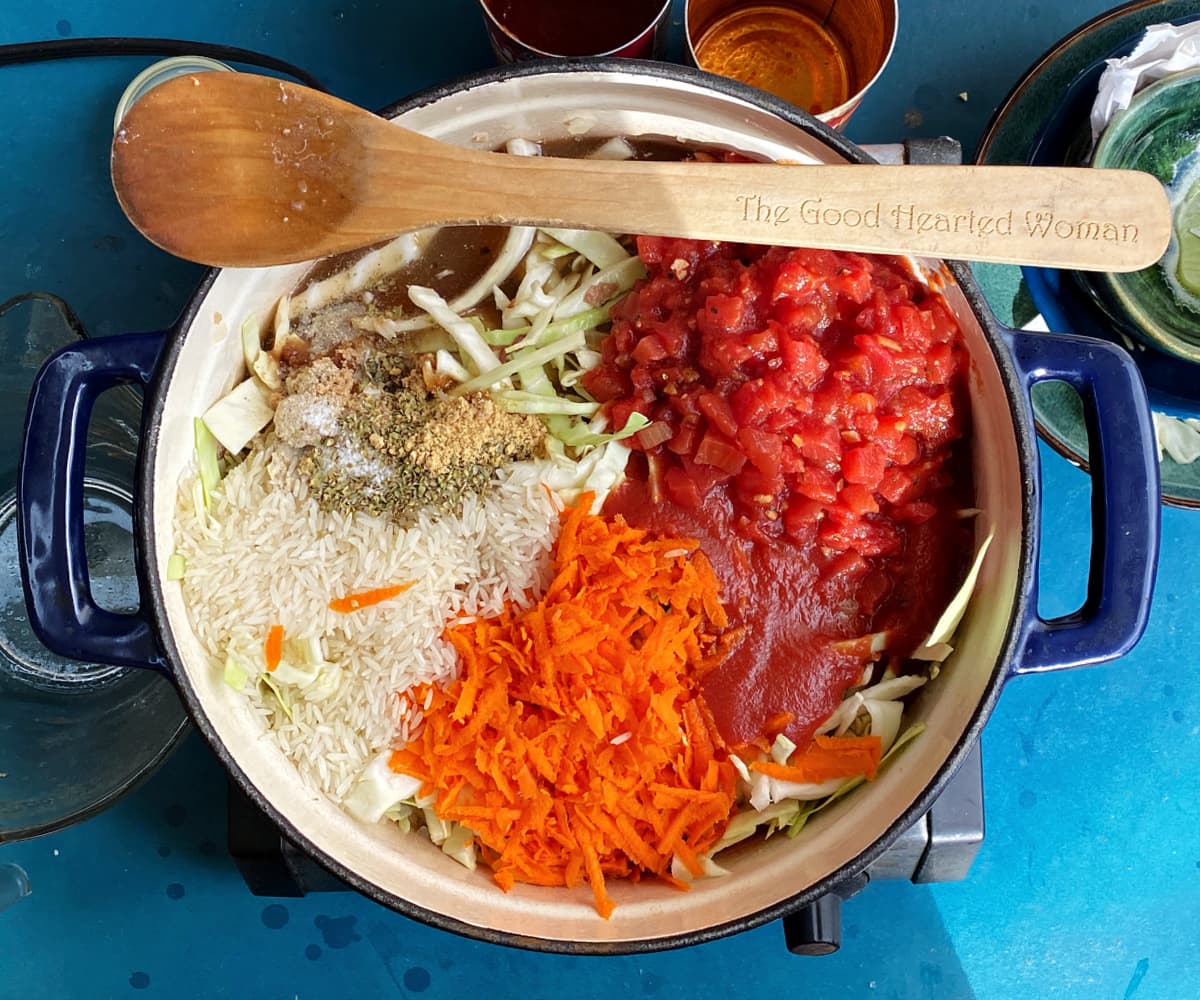 Tomatoes, tomato sauce, carrots, rice, cabbage, and seasonings added to onions and meat in Dutch oven, unmixed. Wooden spoon rests across the top of the pot.