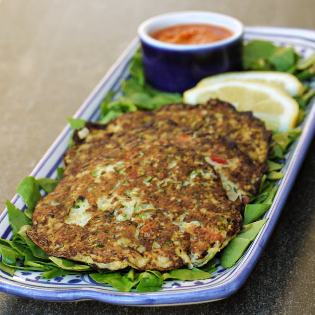 Three zucchini cakes on a bed of greens, with lemon slices and a small ramakin of sauce in the background. 