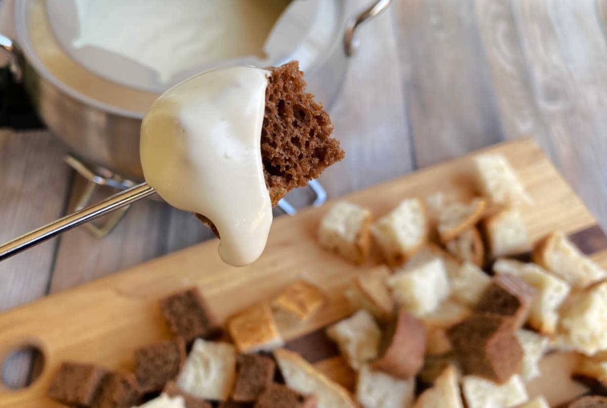 Cube of rye bread on the end of a fondue fork, dripping with melted cheese sauce. Additional bread cubes in the background.