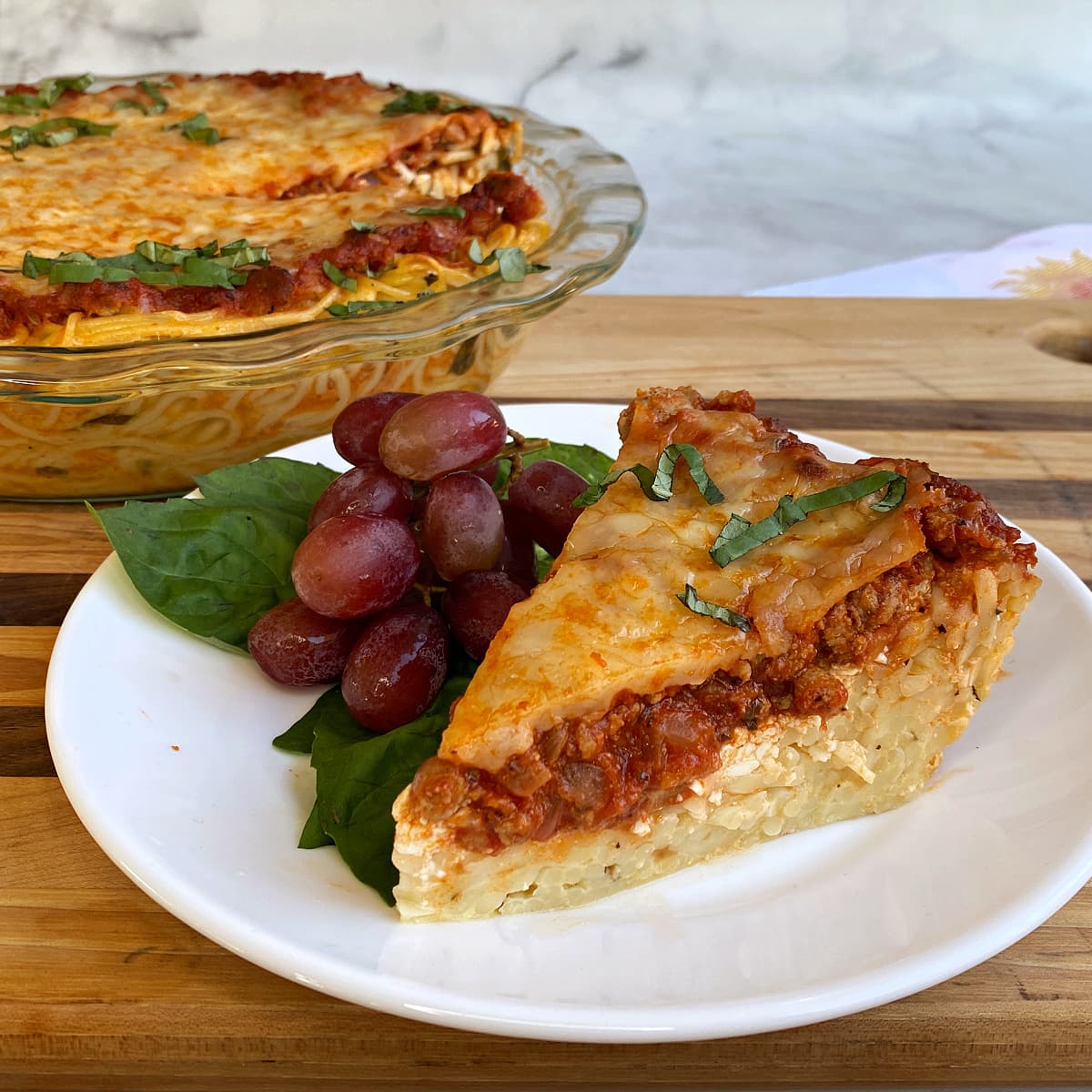 Serving of spaghetti pie cut, on a plate garnished with grapes and fresh basil leaves. 