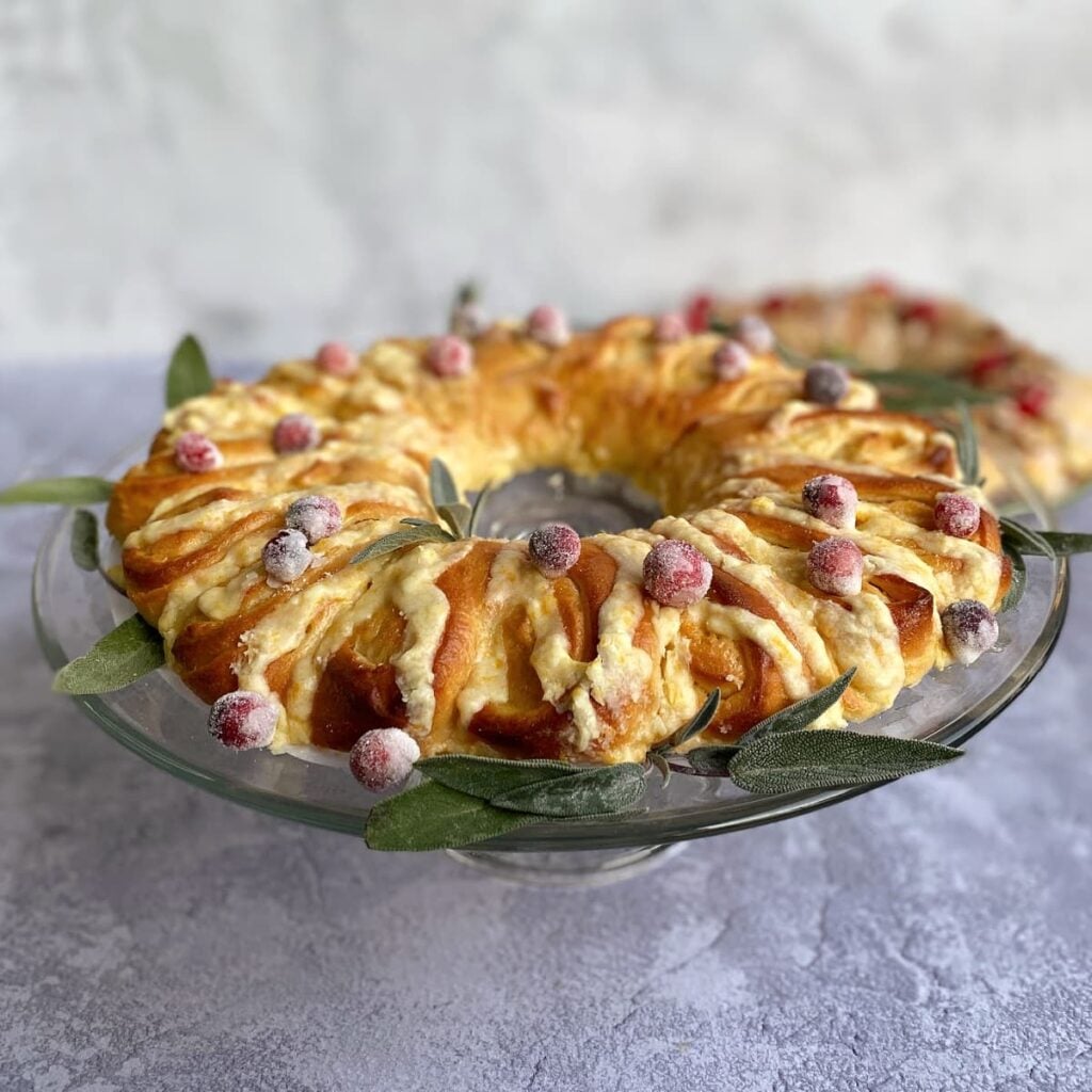 Orange tea ring on a glass cake stand, garnished with small sage leaves and sugared cranberries. Cinnamon tea ring in background.