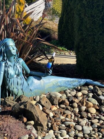 Metal sculpture of a mermaid, resting on a bed of river rocks and holding a bottle of wine.