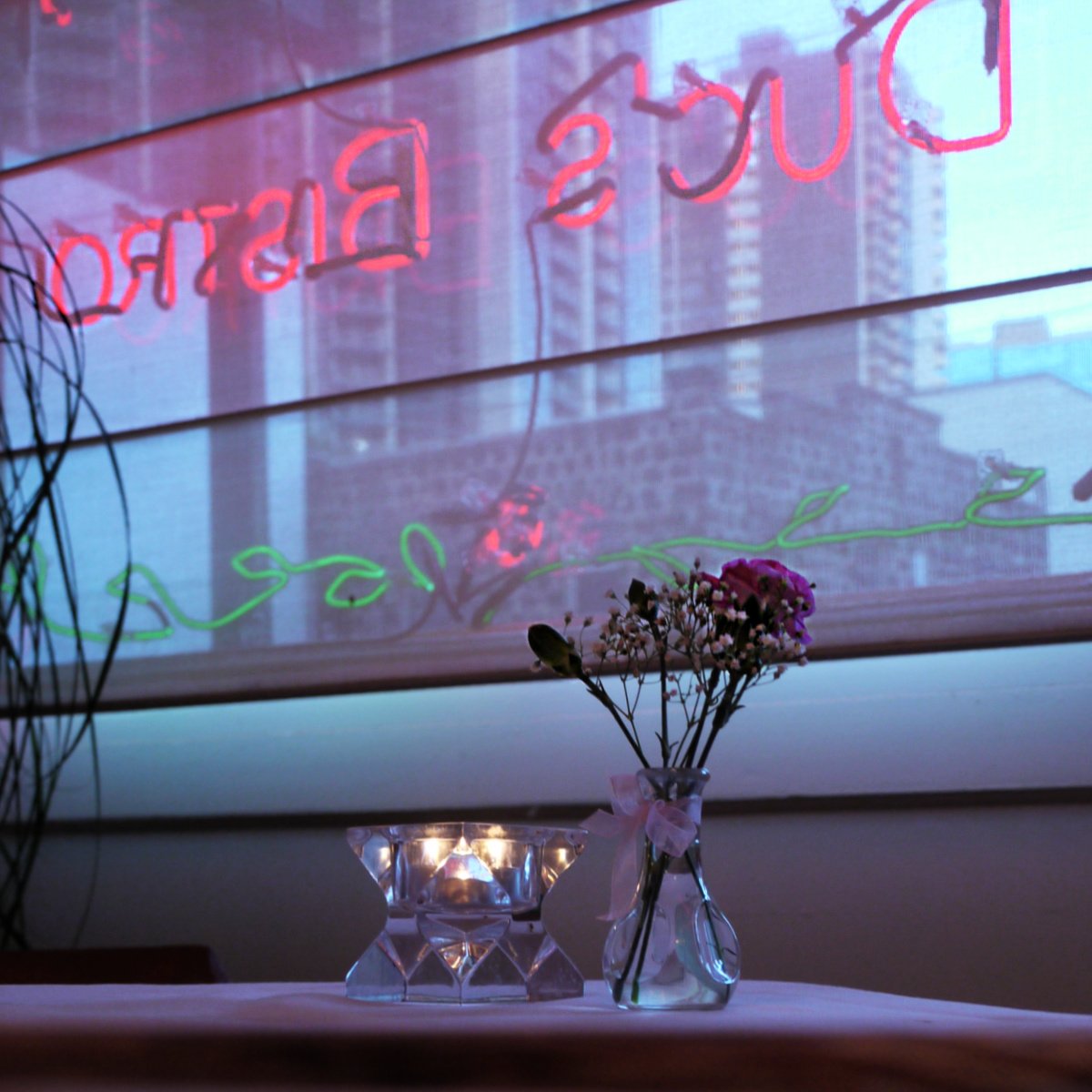 Candle and a small flower vase with a pink carnation and a sprig of baby's breath. A neon sign - Duc's Bistro - read backward through the window. 