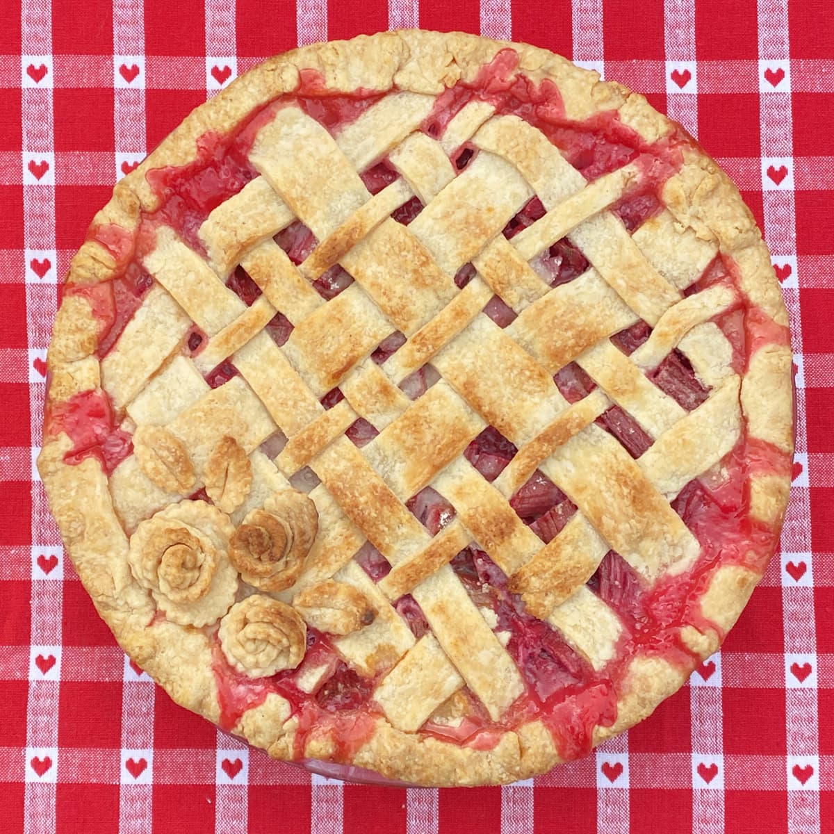 Rhubarb pie with latticed top and pastry roses, setting on a red checked tablecloth. 