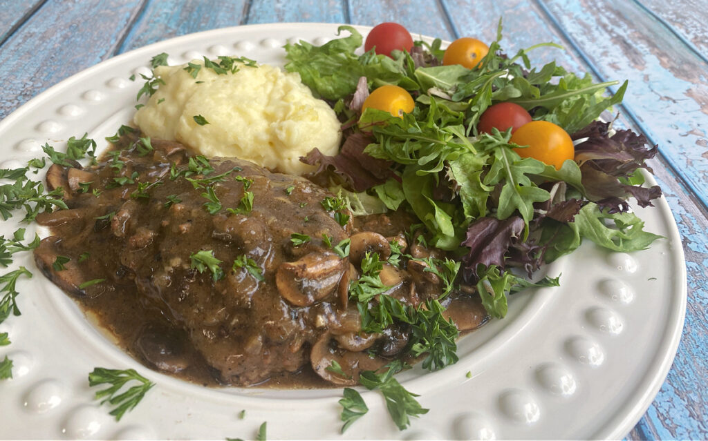 A white plate containing a meal with mushrooms and gravy over meat, accompanied by a serving of mashed potatoes and a side salad with mixed greens and cherry tomatoes. The plate sits on a rustic blue wooden surface.