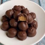 Many peanut butter balls stacked on a plate. One on top has a bite taken out to show center.