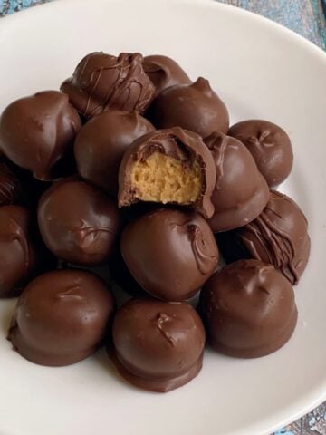 Many peanut butter balls stacked on a plate. One on top has a bite taken out to show center.